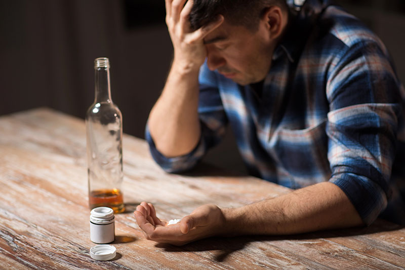 Man with his head in his palm holding pills with a bottle of alcohol on the table